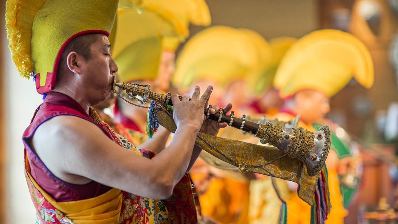 Tibetan monk in red and saffron robe blowing horn instrument